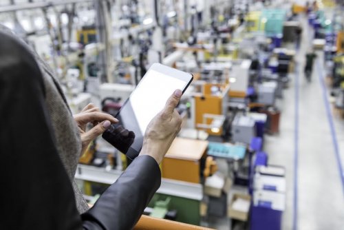 Horizontal color image of businesswoman - unrecognizable person - working with digital tablet in large futuristic factory. Woman standing on top of a balcony, holding touchpad and checking inventory of a manufacturing company on touchscreen tablet. Focus on businesswoman's hands holding black tablet, futuristic machines with incidental person in background.