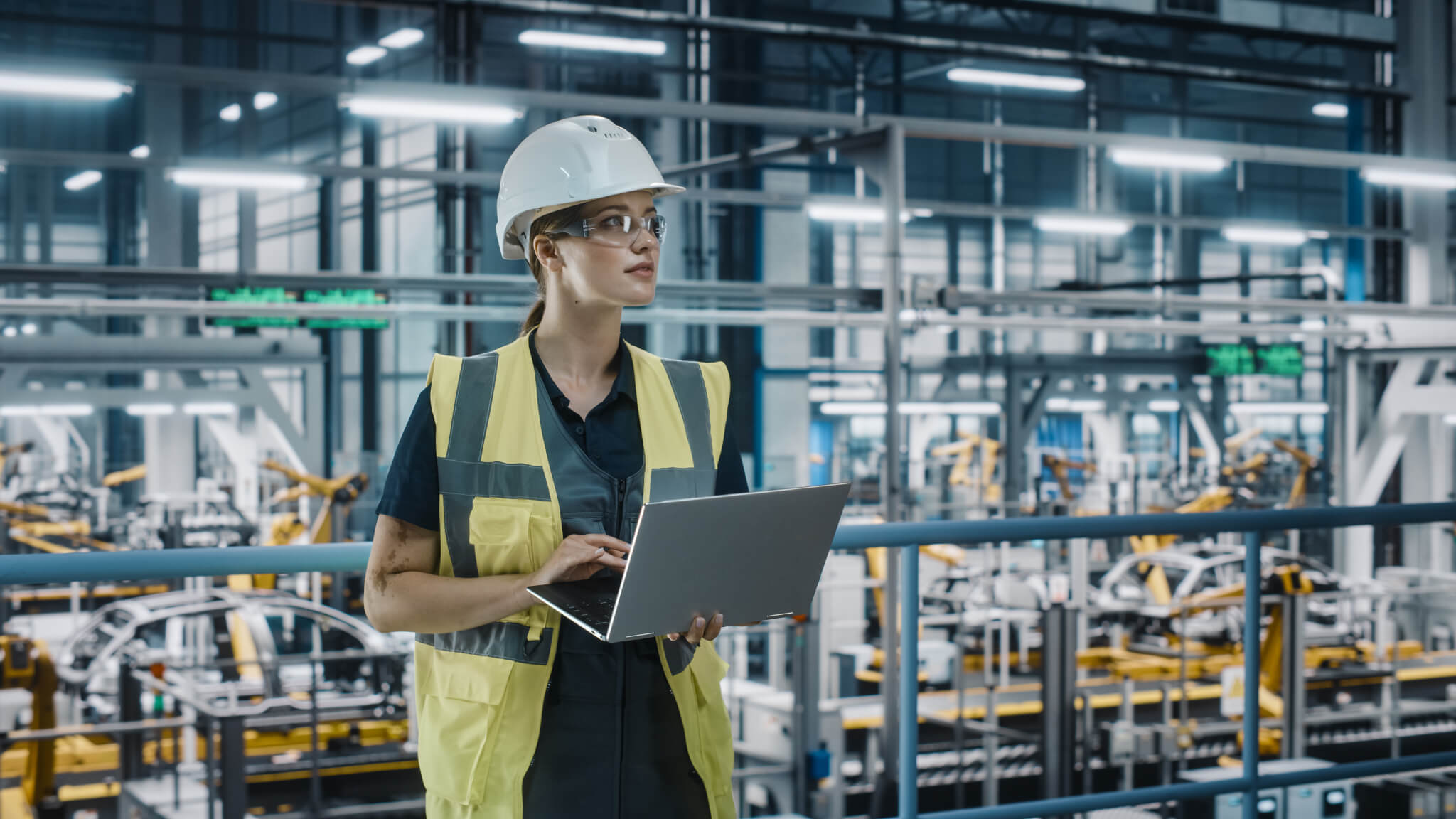 Portrait of Female Automotive Industry 4.0 Engineer in Safety Uniform Using Laptop at Car Factory Facility. Confident Assembly Plant Specialist Working on Manufacturing Modern Electric Vehicles.