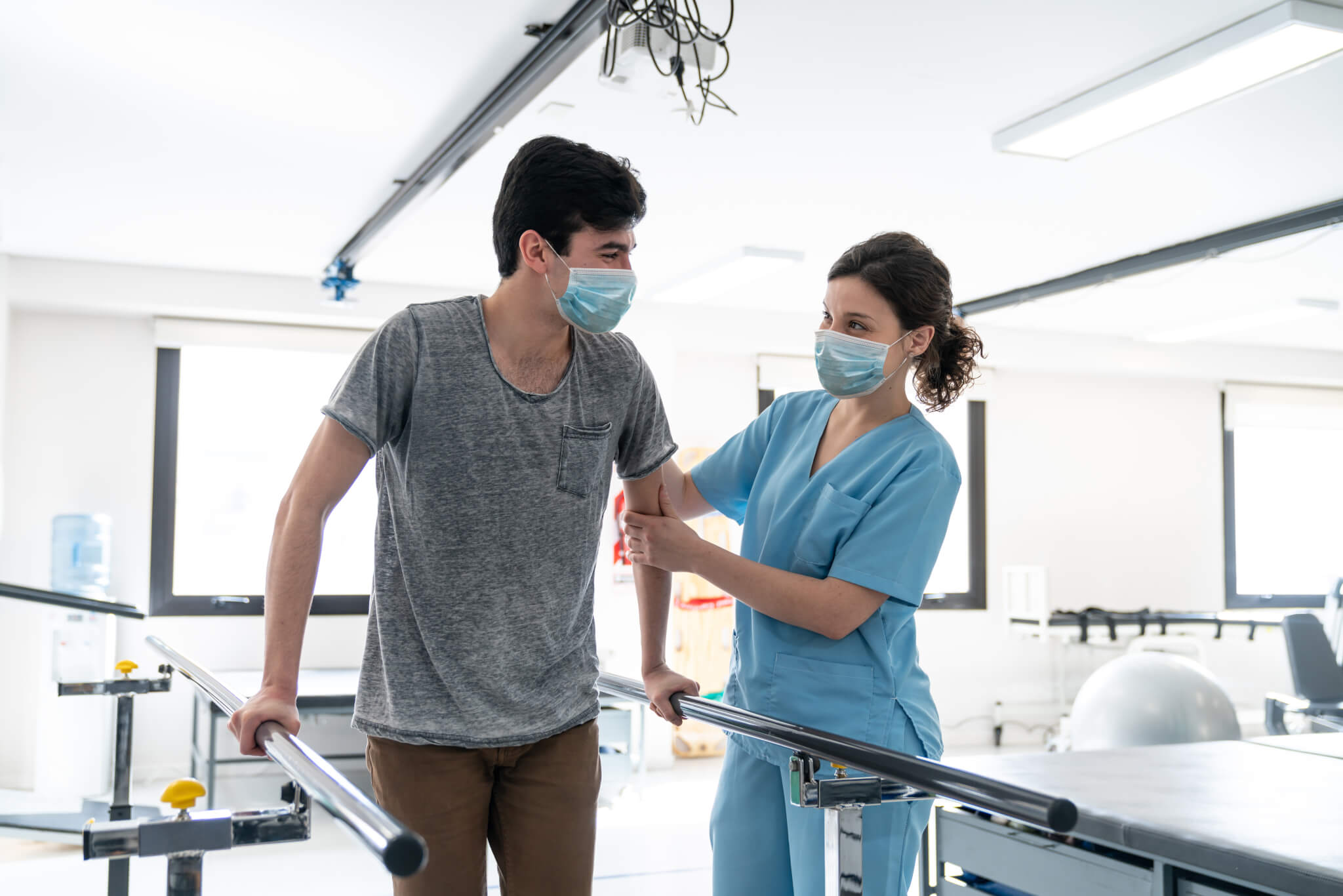 Male patient at physical rehab walking with the help of parallel bars while therapist supports him both wearing protective face masks